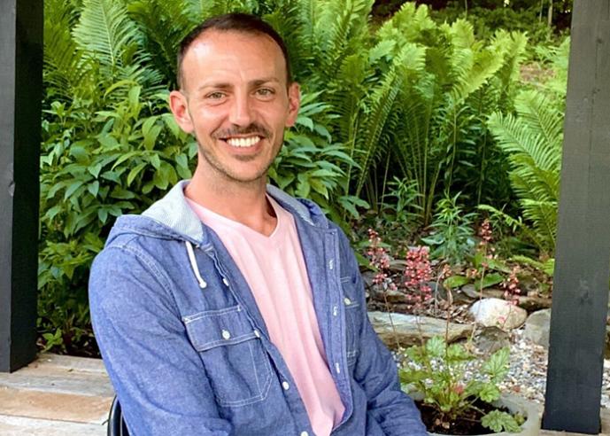 A man casually dressed, smiling, looking at camera, with green plants in background.