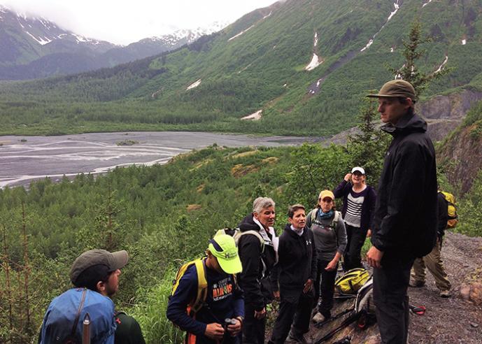 A group of hikers stop to listen to a fellow hiker speak, with water and mountains in background.