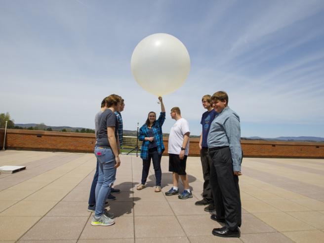 A group of students stand on an observation deck, a female student holds a weather balloon.