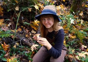 A young woman sitting on a log, wearing a hat, holds a mushroom in the woods, ferns and leaves in background.