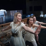 A group of female actors wearing white perform on a stage.