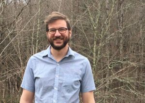 A man with glasses and a beard, smiling, looking at camera, with trees in background.