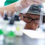 A student wearing a hat and glasses works in a lab.
