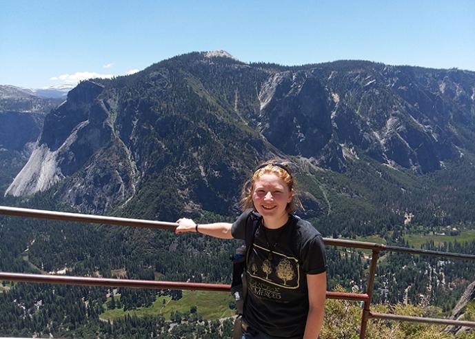 A young woman stands in front of a mountain scenic overlook, smiling, looking at camera.