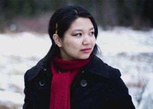 A young woman with long, dark hair, a red scarf and a pencil behind her ear, stands in a snowy exterior, looking off to her left side.