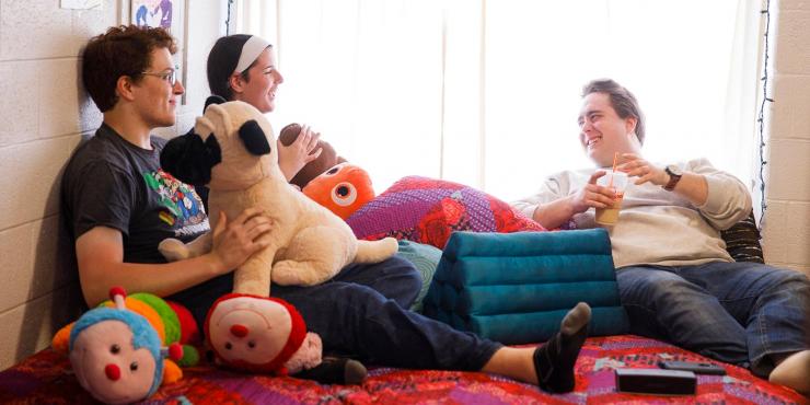 Three friends hang out in a dorm room, sitting on a bed with stuffed animals and pillows.