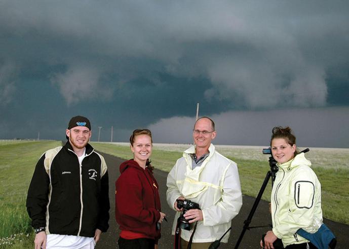 Four people stand in a road wearing rain gear, smiling at camera, with storm clouds in background.