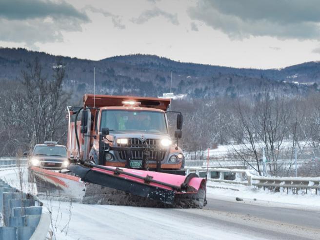 A large snowplow removes snow on a road, a pickup truck follows, trees and hills in the background.
