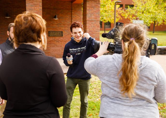 A young man holding a mircophone, smiling, with a camera crew with equipment in foreground.