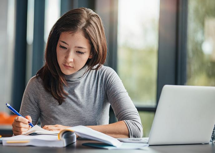 A female student with long, brown hair, sits at a desk and holds a pen, studying.