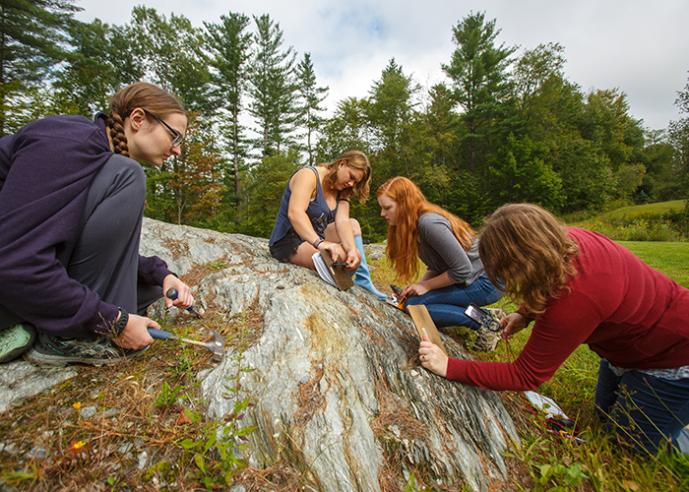 Four female Environmental Science students sitting on a large rock, studying, taking notes, with trees in background.
