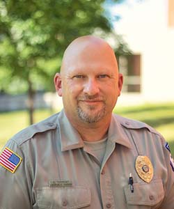 A bald man with a blond goatee smiles at the camera. He is wearing a tan suit with a badge. 