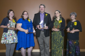 Group of men and women dressed nicely against a brown backdrop smiling at the camera while holding awards.