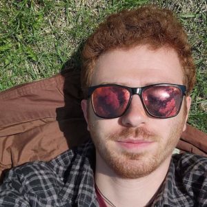 Young man with red hair wearing sunglasses looks into the camera.