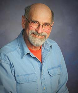A balding man with glasses and a while beard smiling at the camera. He is wearing a blue button up shirt.