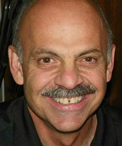 A close up of a man's face. He is bald with a graying mustache and he has a very large smile on his face.
