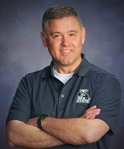 A man with gray hair smiling at the camera with his arms crossed over his chest. His shirt is a gray polo.