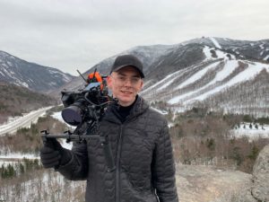 A young man in black, wearing a black hat holds a video camera. There is a snowy ski area in the background.