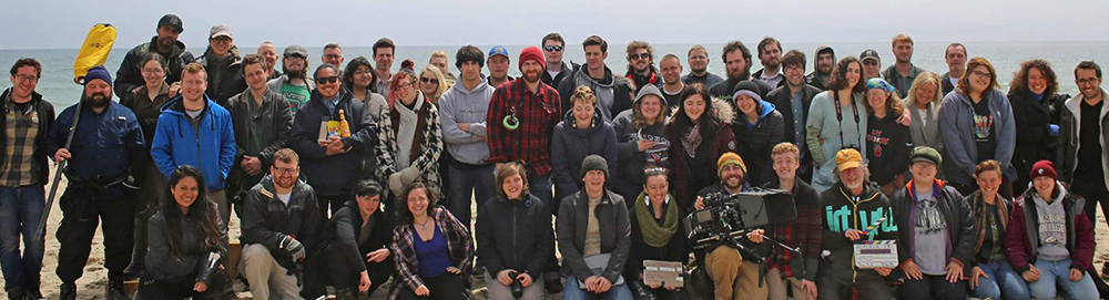 A large group of people of all ages smiling at the camera. They are on a beach, the ocean is in the background.