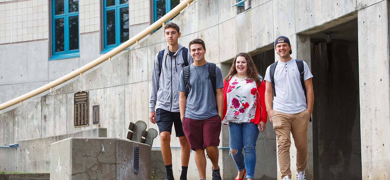 For students, three guys and one girl, are smiling and walking towards the camera.