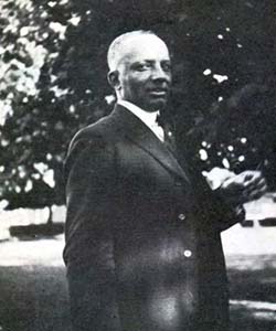 A black and white side view of a man as he looks at the camera, he is wearing a suit and tie.