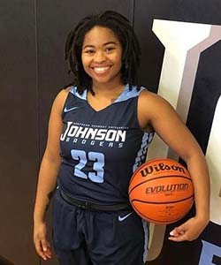 A young woman with dreadlocks smiles at the camera. She is wearing a Johnson Badgers jersey and holding a basketball.