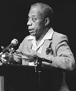 A black and white photo of a Black man standing at a podium talking into a microphone. He is wearing a suit and has short hair.