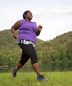 A Black woman wearing a purple shirt and black leggings running on grass with green hills in the background.