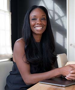 A black woman with long black hair sits with her arms rested against the desk, she's smiling at the camera.