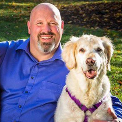 A bald man with a gray beard wears a blue button up shirt, has his arm wrapped around a golden retriever dog, and smiles at the camera.