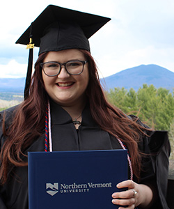 A girl with brown hair in graduation regalia holding a diploma against a mountain background.