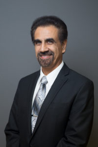 A main with dark hair and a goatee smiles at the camera while wearing a suit and tie.