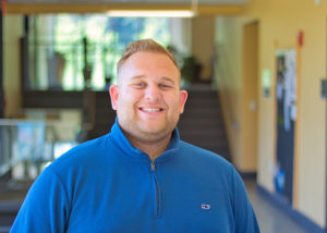 A young man with short blond hair in a blue quarter zip shirt smiles at the camera.