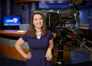 A girl with long dark hair smiles at the camera. She is wearing a purple dress and leaning against a giant video camera, hand on her hip. She is in a news studio.