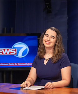 A dark haired girl sits in a news studio behind a desk. She is wearing a dress and smiling at the camera with her head tilted. A screen behind her says "news7"
