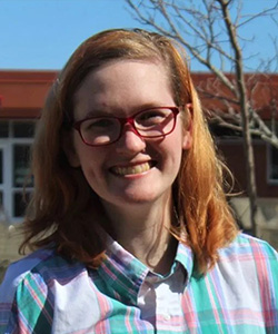 A strawberry blond girl in red glasses smiles at the camera. She's standing outside wearing a plaid button up shirt.