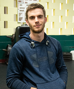 A young man with short hair and a beard wears a dark blue hoodie and smirks at the camera.