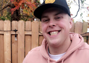 A young man in a black baseball cap smiles brightly at the camera. He's wearing a pink hoodie.