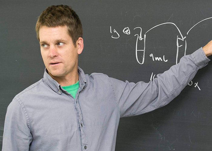 A photo of short haired man standing against a blackboard pointing at chalk marks.