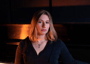 A young woman with long blond hair sits in a dark room at a piano staring into the camera.