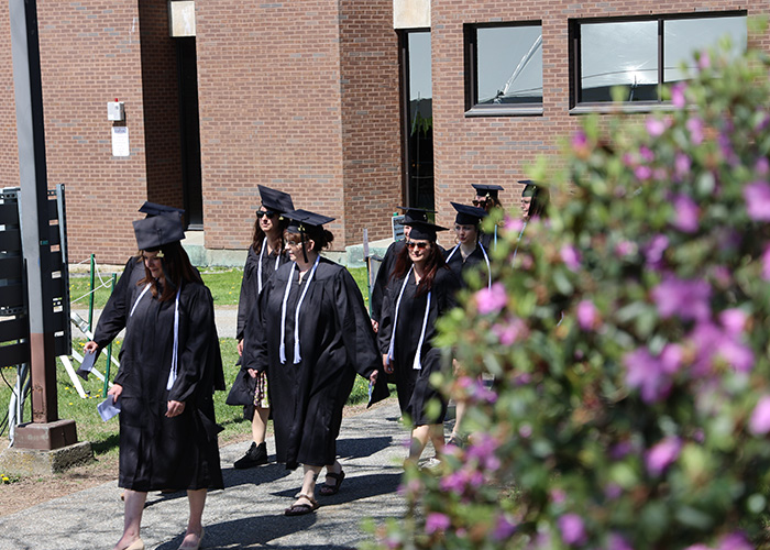 A group of people in graduation regalia walking past a bush of purple flowers.