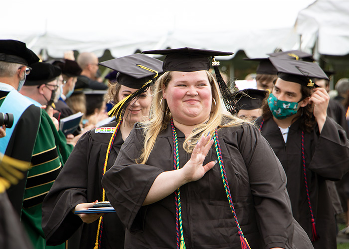 A woman with long blond hair is waving and smiling while wearing graduation regalia.