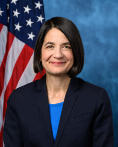 A headshot of a woman with short dark hair smiling at the camera, American flag in the background.